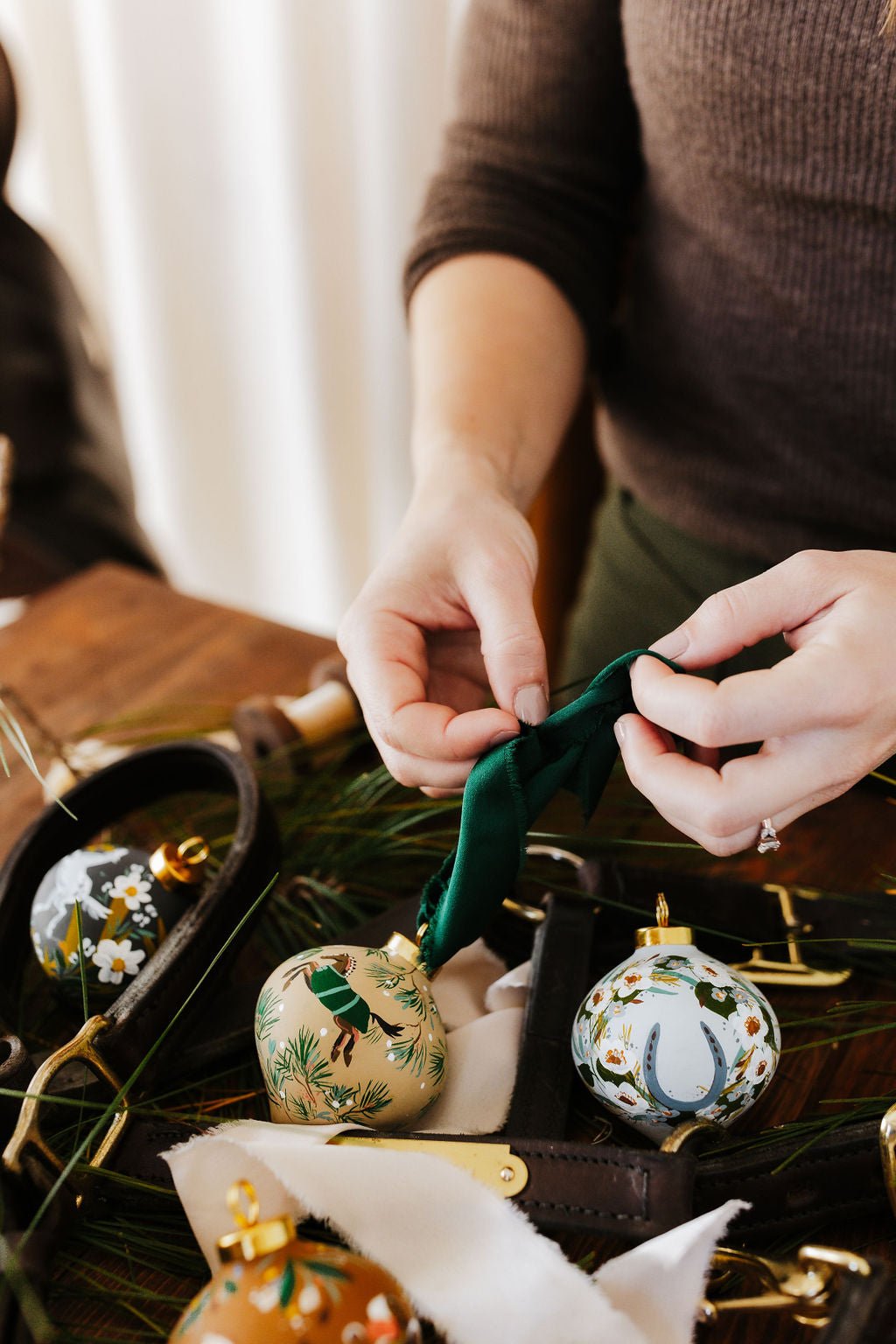 Artist Larissa Ann decorating equestrian Christmas ornaments with green ribbon on a wooden table.
