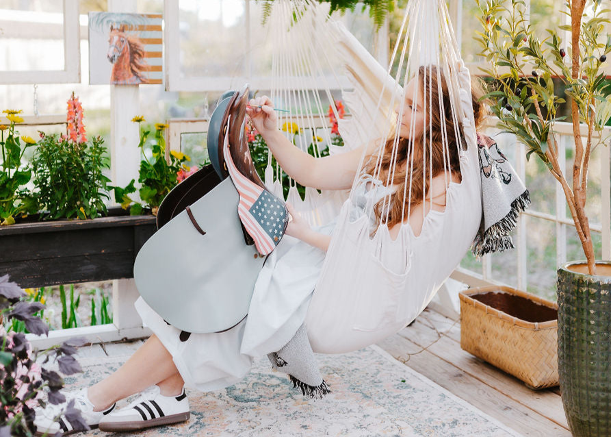 Equestrian artist Larissa Ann paints a saddle while sitting in a hammock on a patio with plants and flowers around