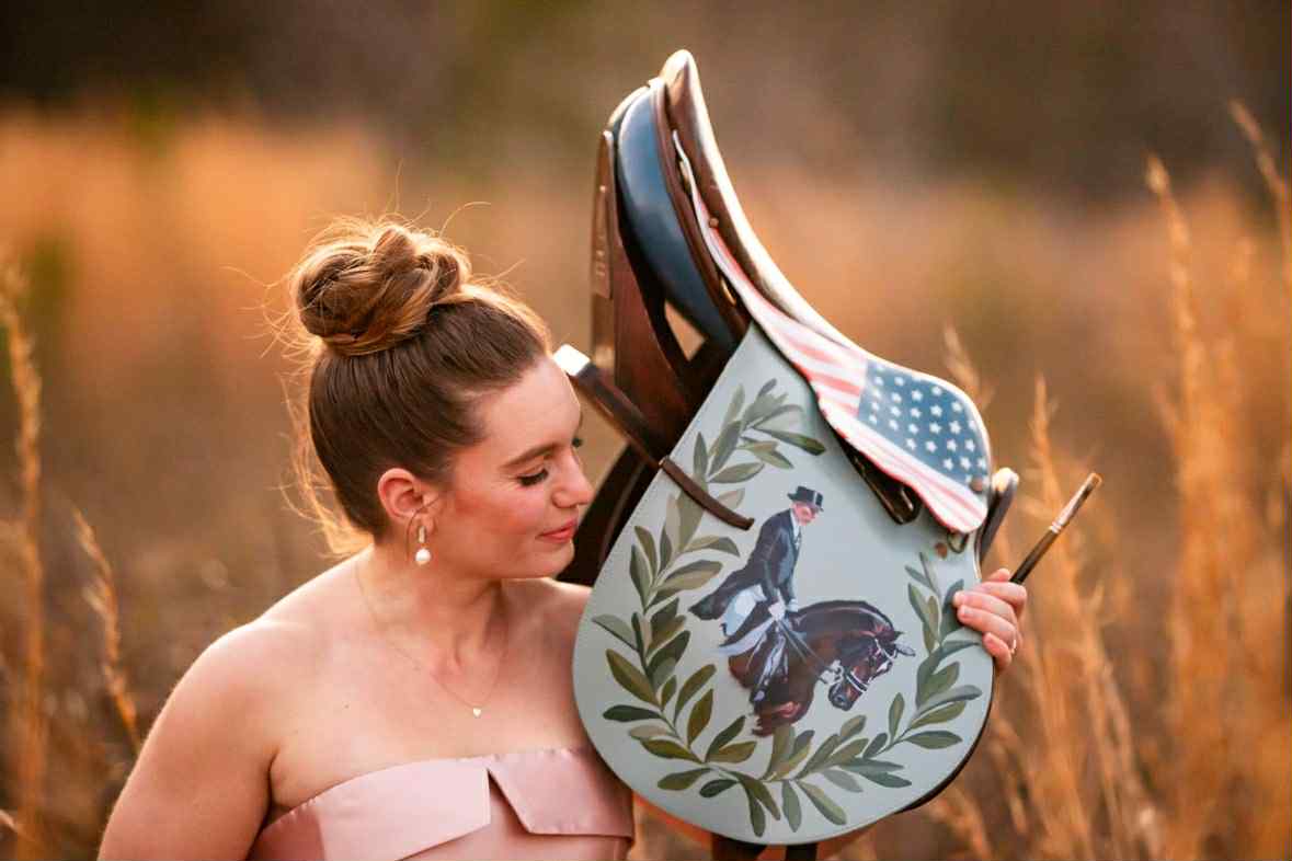 Equestrian artist Larissa Ann holding a custom painted horse saddle in a field