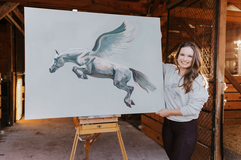 Equestrian artist Larissa Ann holding a painting of a pegasus in a horse stable