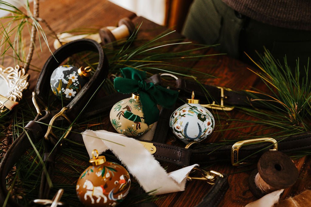Decorative hand painted equestrian Christmas ornaments on a rustic wooden surface with greenery.
