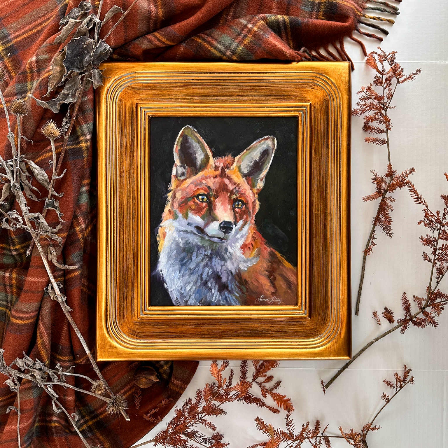 Framed painting of a fox on a surface with plaid fabric and branches.