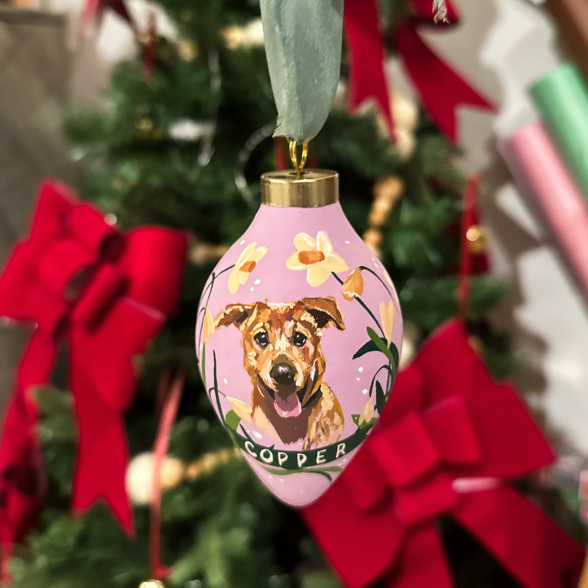 A pink Christmas ornament featuring a painted dog portrait and the name 'COPPER' on a ribbon, hanging from a green ribbon against a decorated Christmas tree with red bows.