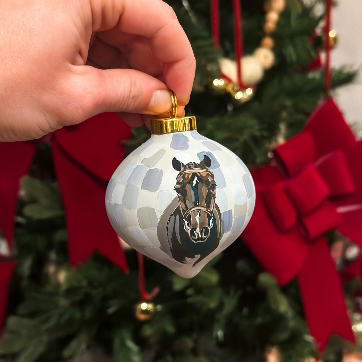 A hand holds a white Christmas ornament featuring a painted horse portrait, with a blurred Christmas tree and red bows in the background.