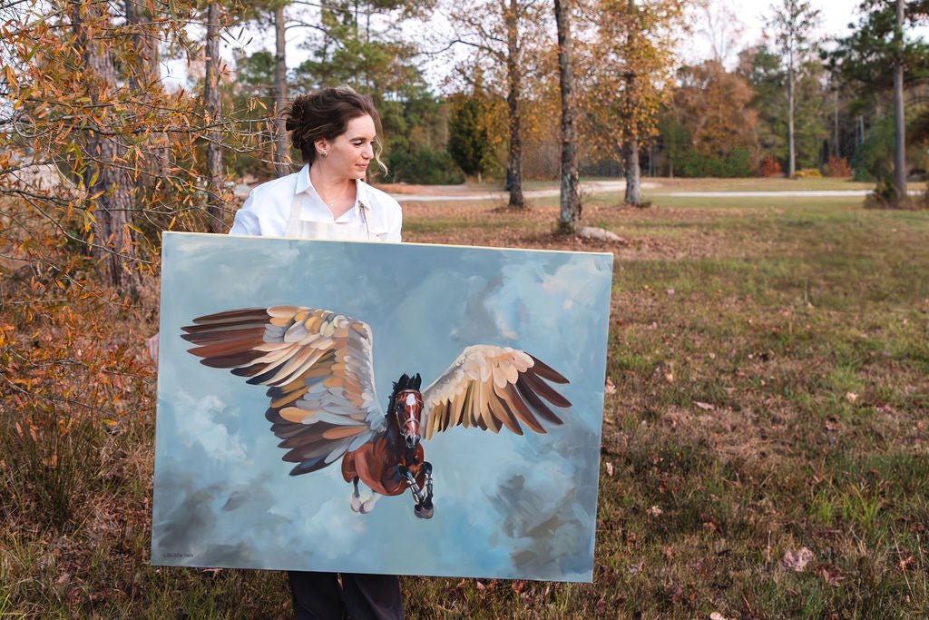 Artist Larissa Ann holding a painting of a horse with wings in an outdoor setting