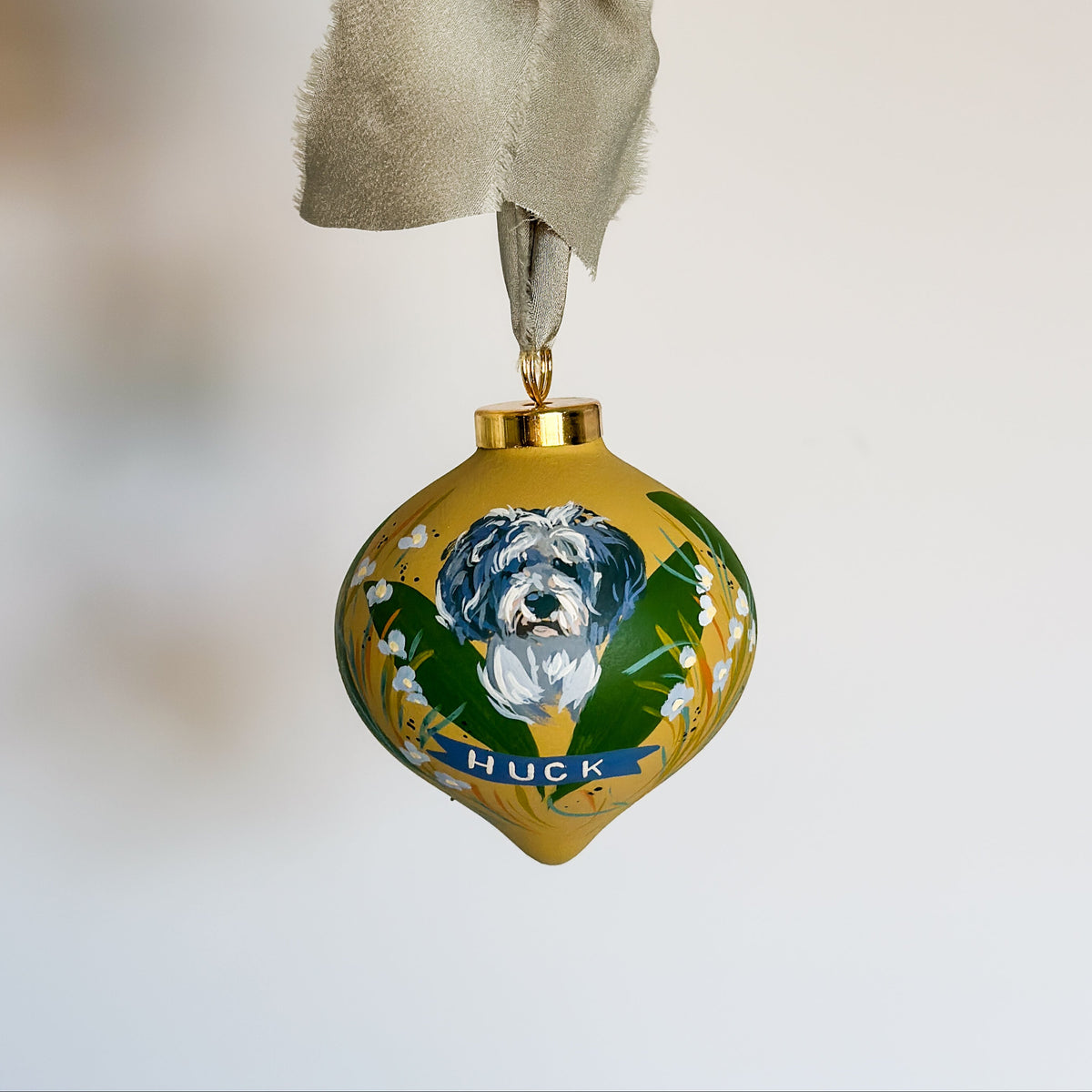 custom hand-painted ornament with a pet dog and his name. The ornament is gold with a painted dog portrait, green leaves, white flowers, and a blue banner reading "HUCK," attached to a frayed silk ribbon. thoughtful heirloom gift