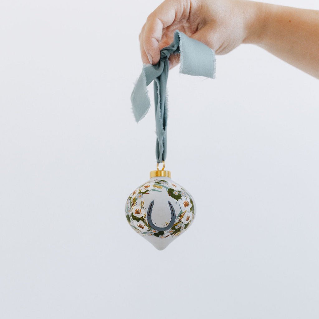 A decorative equestrian ornament with a hand painted horseshoe wreath and blue ribbon against a light background