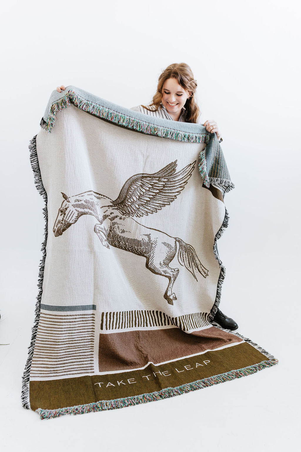 Artist Larissa Ann holding atapestry blanket with a Pegasus design and 'Take the Leap' text on a white background