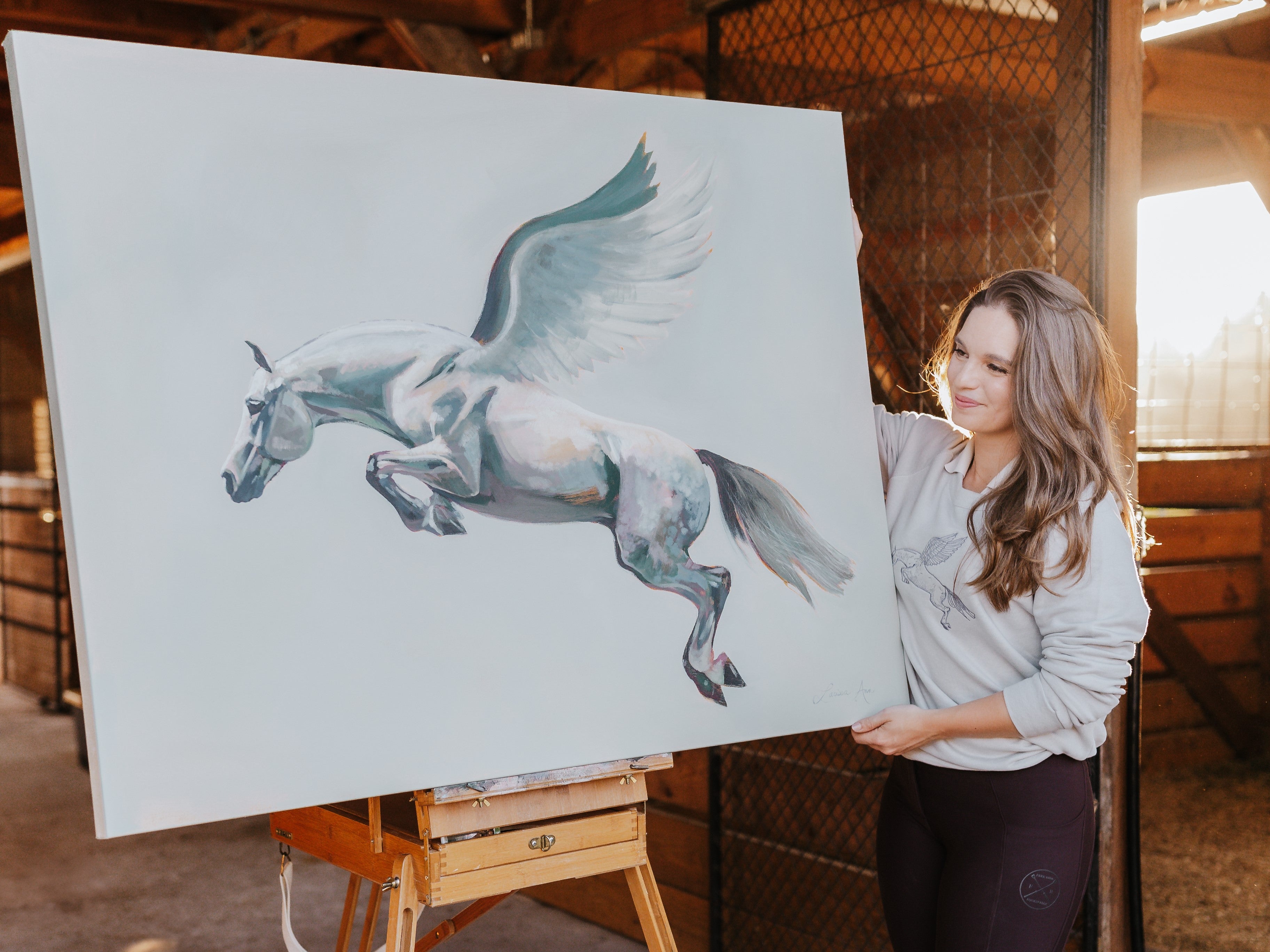 Equestrian artist Larissa Ann holding an original painting of a winged horse in a rustic barn setting
