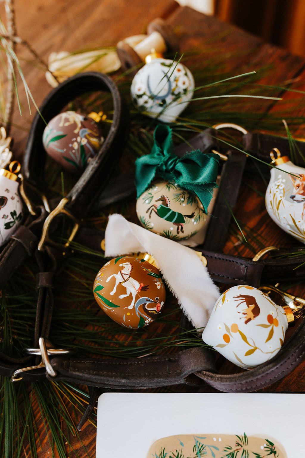 Hand painted equestrian decorative Christmas ornaments with horse designs on a wooden surface with greenery