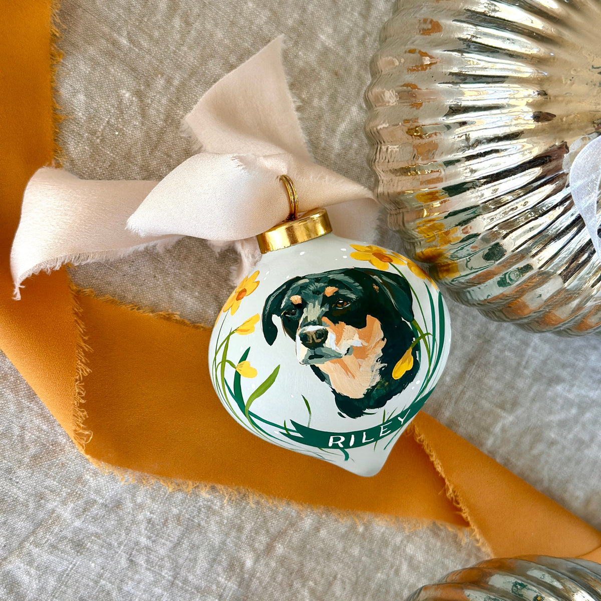 A close-up of a custom painted Christmas ornament shaped like a teardrop, featuring a painted black, white, and tan dog portrait with green and yellow floral accents and the name 'RILEY' at the bottom. The ornament is tied with a white satin ribbon and placed on a textured cream fabric with orange ribbon and silver ribbed holiday decor nearby.