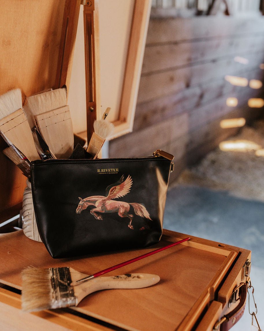 Custom painted black leather clutch with a winged horse design on a wooden easel with paintbrushes