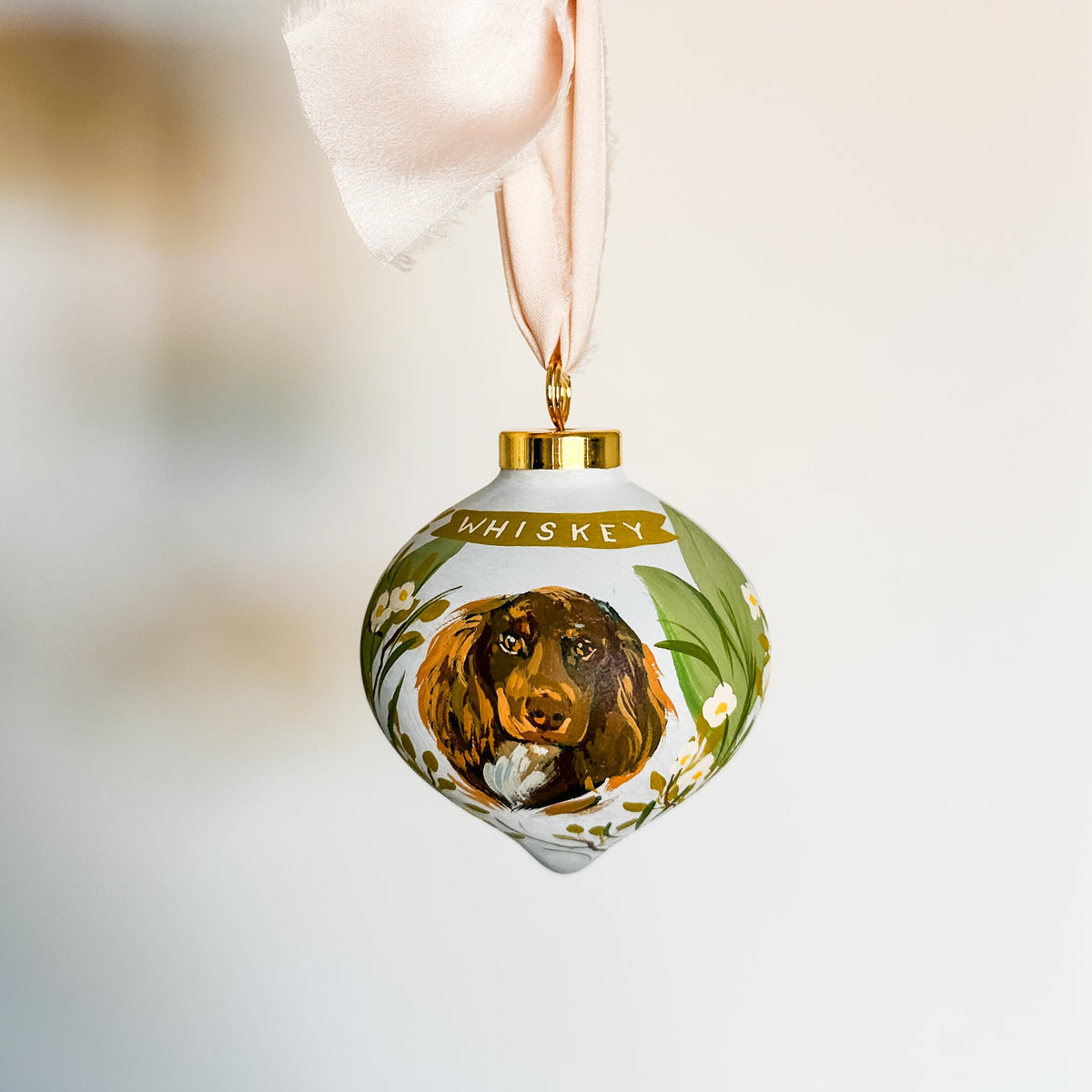 custom pet portrait Christmas ornament shaped like a teardrop. The ornament is white with a gold cap, a silk cream ribbon, and a painted illustration of a brown long-haired dog with green leaves and small white flowers. bespoke commissioned unique gift for someone who has everything